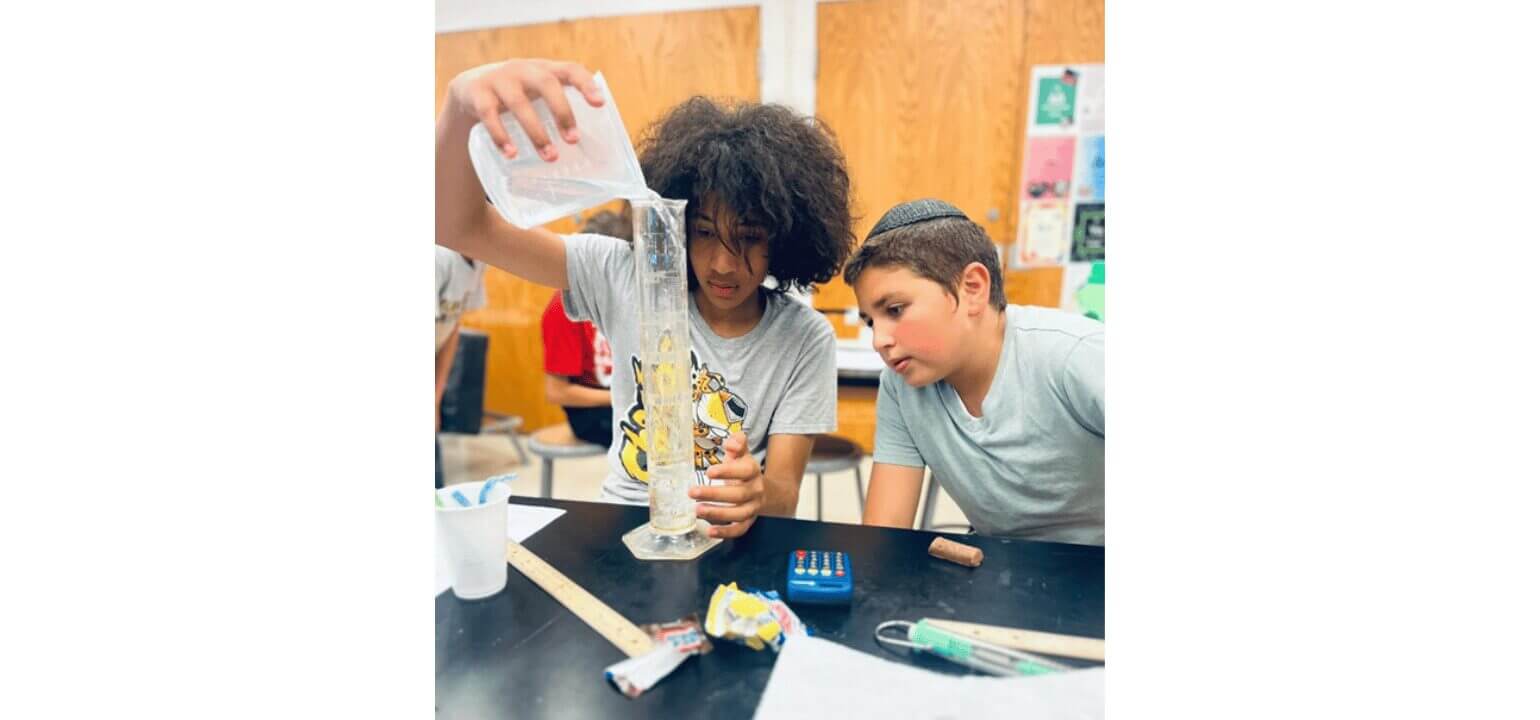 Two boys in a classroom conducting a science experiment, pouring and measuring liquid in a graduated cylinder.