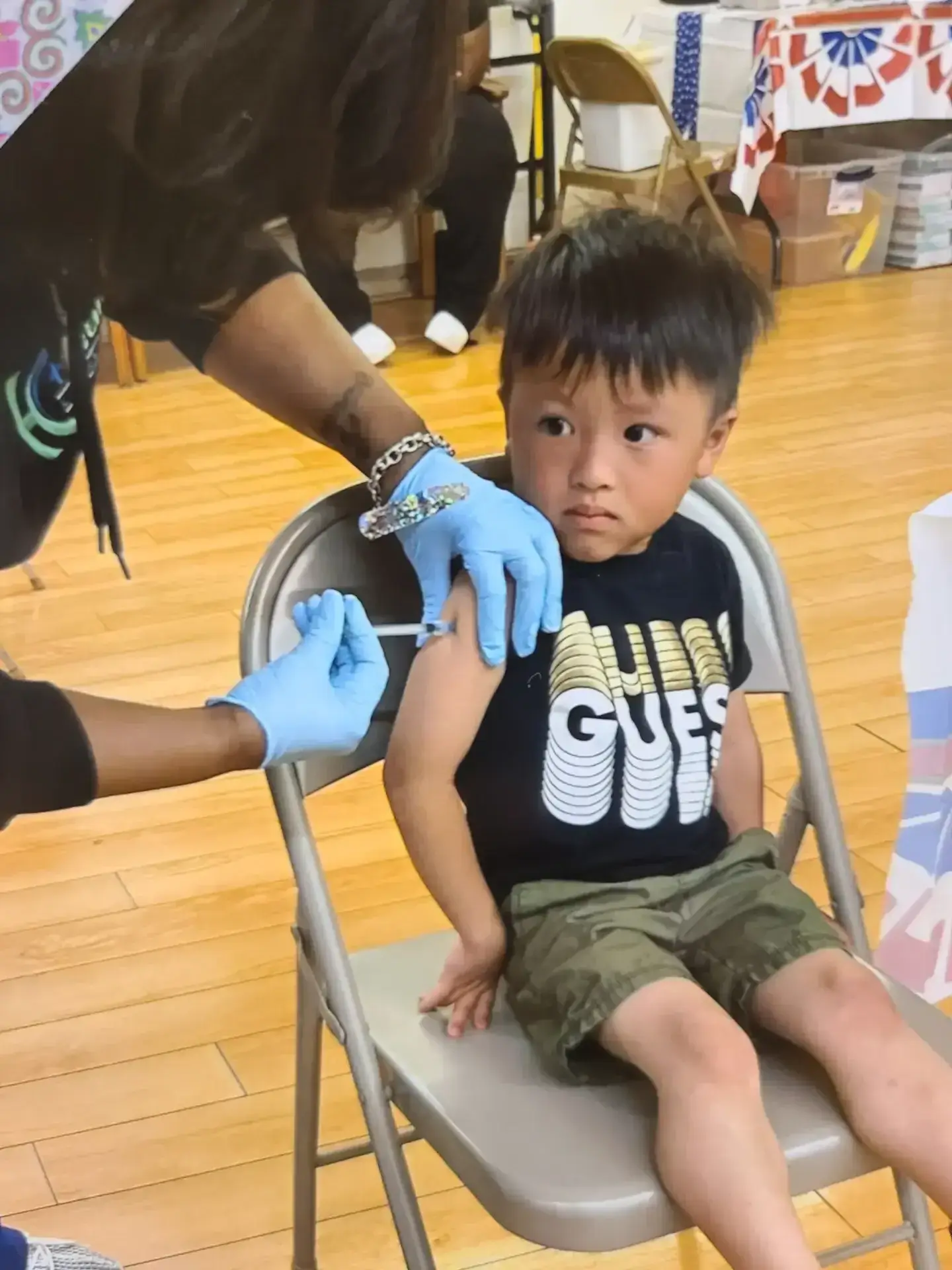 Young Hmong American boy receives vaccine shot from a gloved healthcare worker at a 2023 community health event.