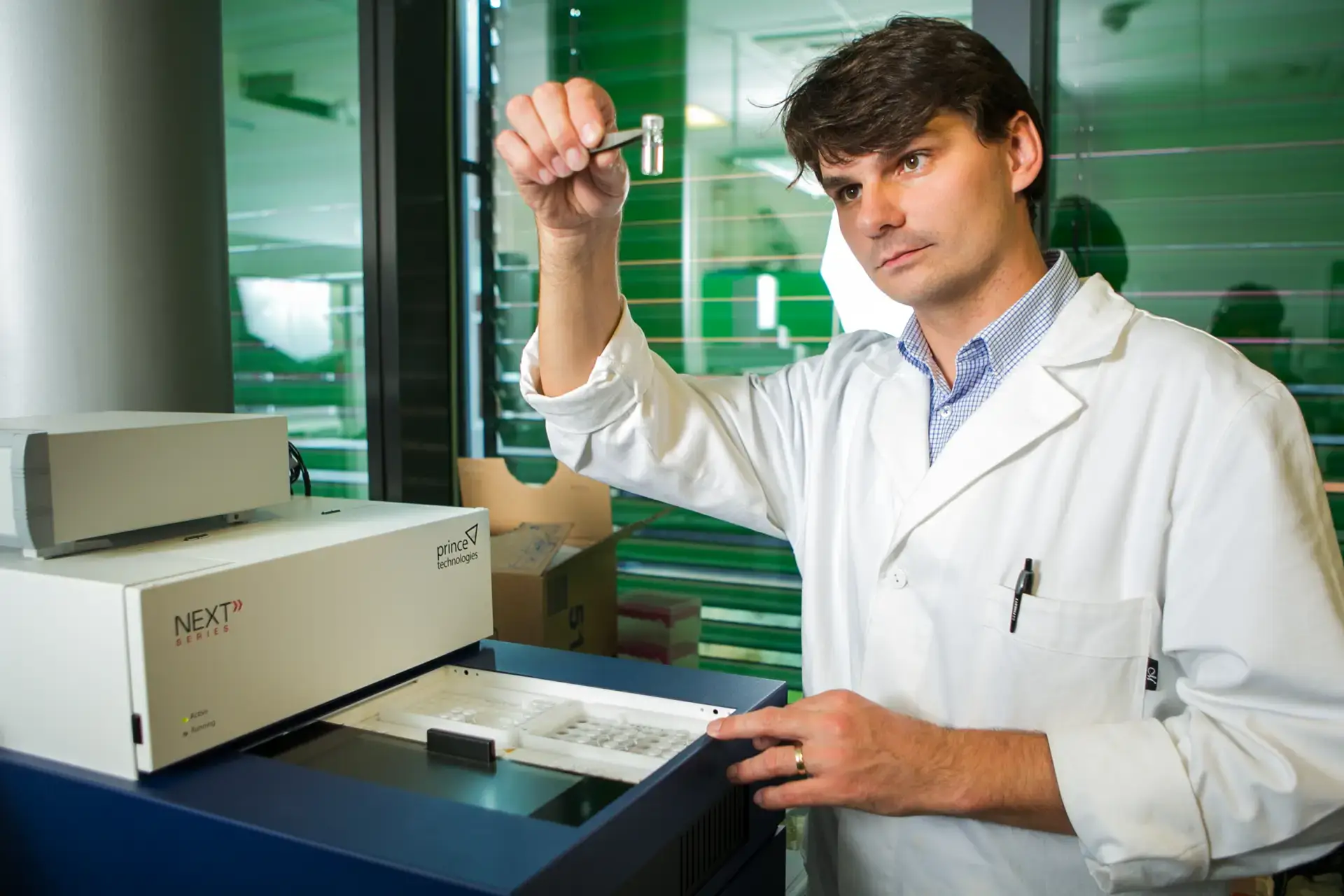 Masaryk University chemistry scientist in lab coat examines sample vial with tweezers. Prince Technologies lab instrument.