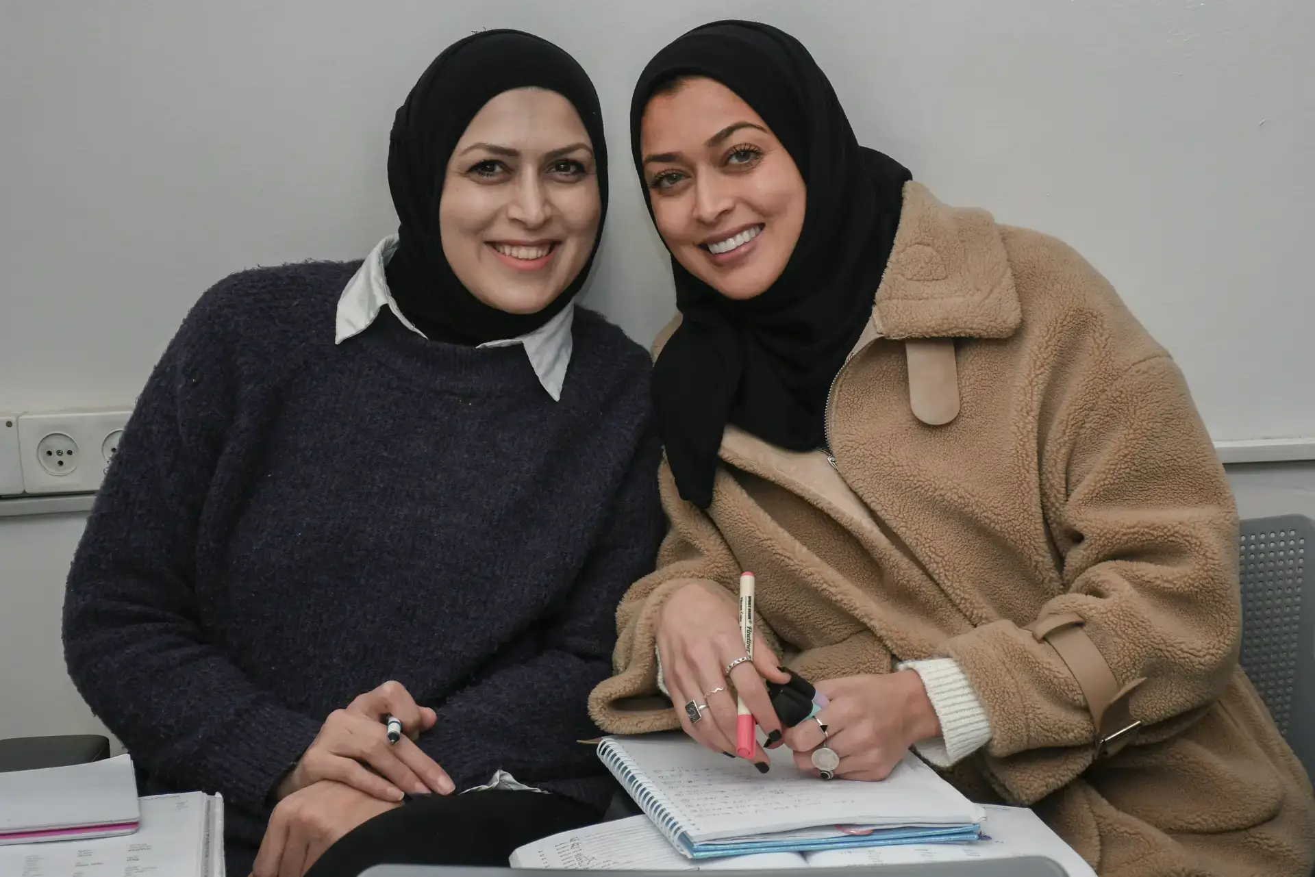 Two women in hijabs smiling, engaged in a Lissan social equality program with notebooks.