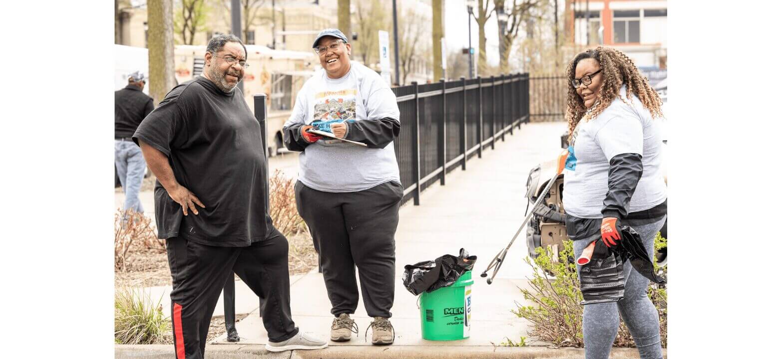 Three diverse volunteers smiling during an outdoor community cleanup, holding a grabber tool and green bucket.