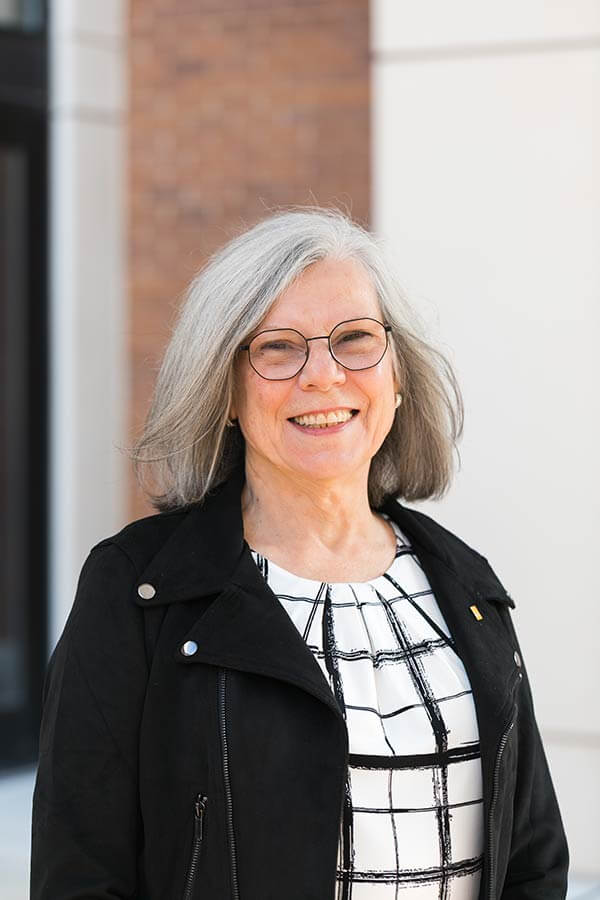 Marg Foster smiling in professional headshot with short brown hair, glasses, and a black jacket over a patterned blouse.