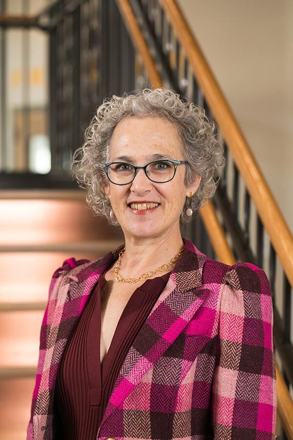 Michelle Berrong Bader smiling, wearing teal glasses and a colorful patterned jacket, against a teal background.