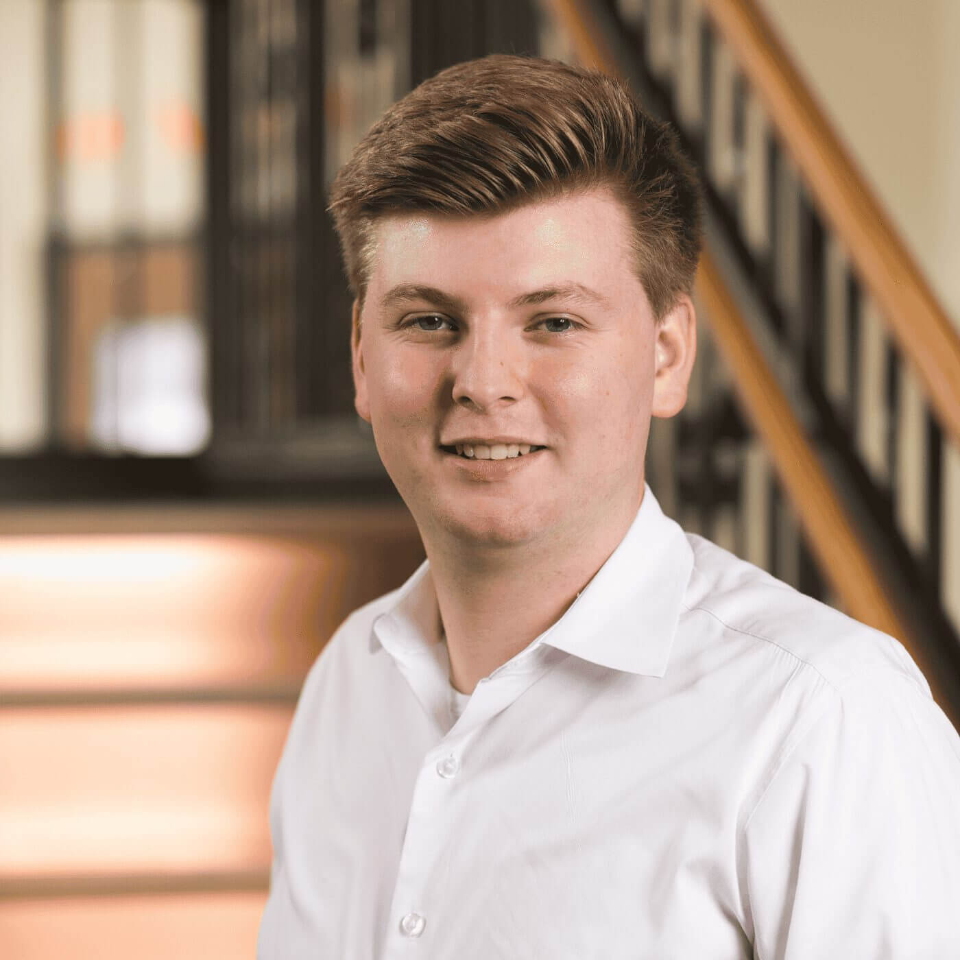 Sam, a smiling young man in a white collared shirt, stands in a modern office with a staircase backdrop.