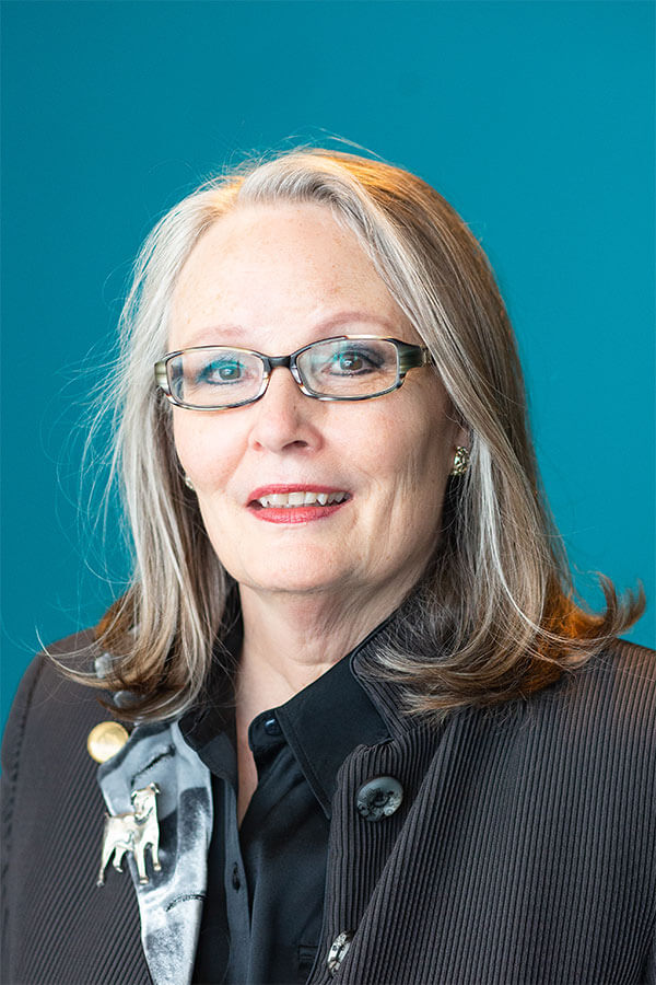 Deirdre Britt, a woman with graying hair and glasses, smiles in a professional headshot, wearing a dark jacket and dog brooch.