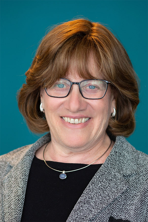 Francie Wolff smiling, wearing black glasses, a herringbone blazer, and a necklace against a teal background.