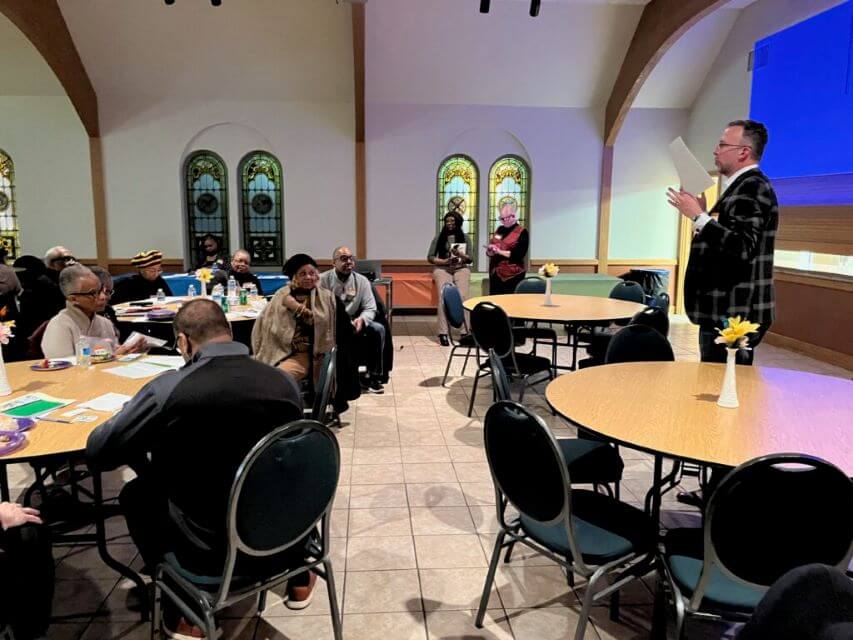 Engaged Community Gathering with Speaker Speaker presenting to a diverse community group gathered at tables in a large room with stained glass windows.