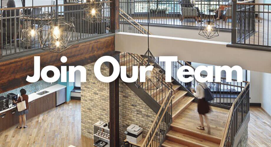 Modern, open-plan office interior with industrial accents. A person on stairs, another at a kitchenette. 'Join Our Team' displayed.