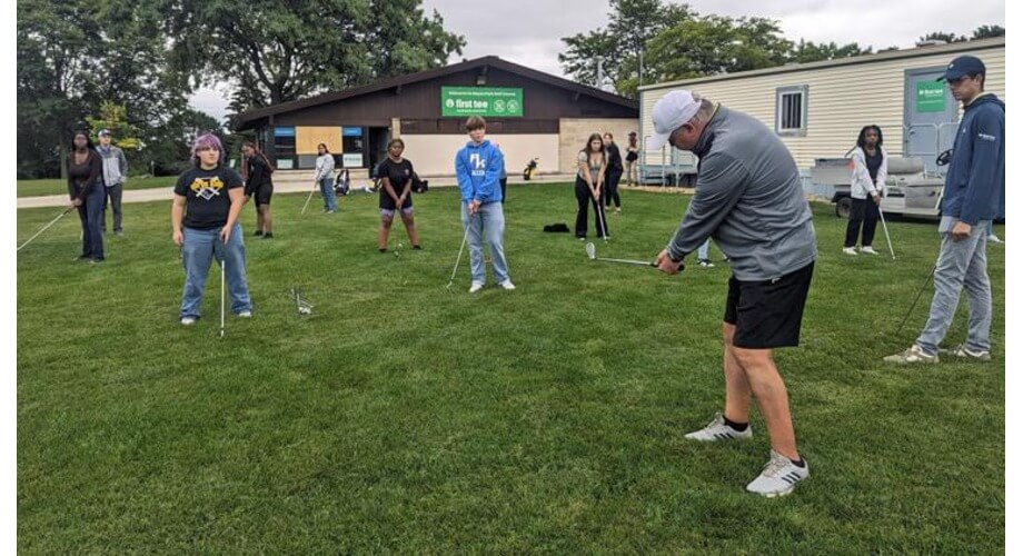 Diverse students learn golf techniques from an instructor outdoors at a First Tee program. Focus on youth skill development.