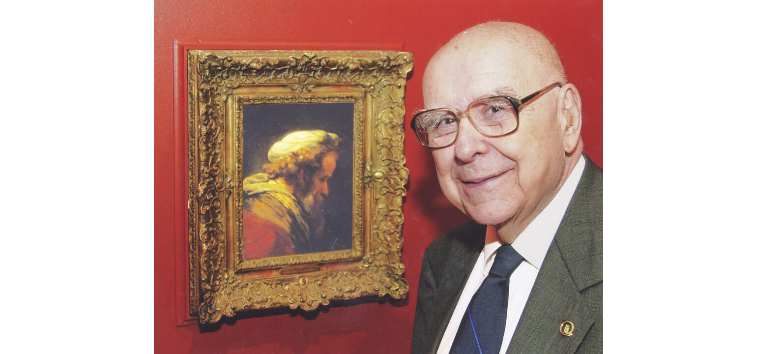 Chemist and art collector Alfred Bader smiles next to a framed Rembrandt portrait on a red wall.