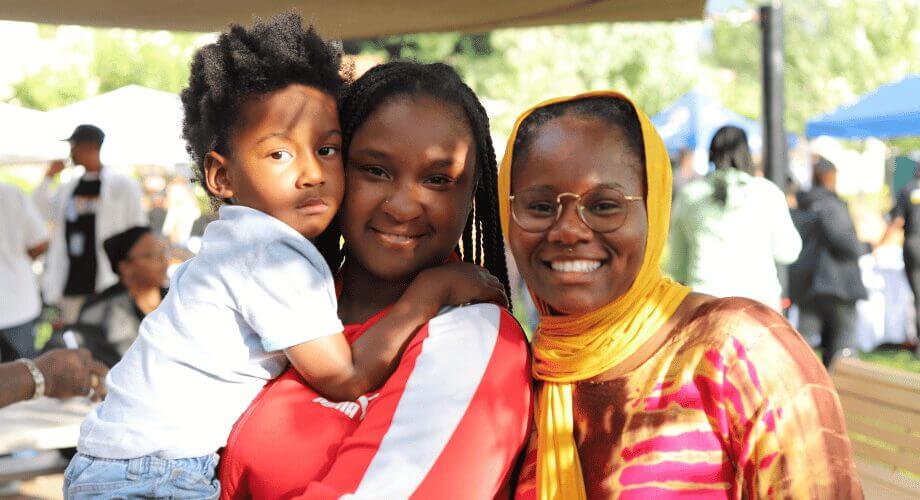 A joyful African American boy, a woman with braids, and a woman in a yellow hijab smiling at Bader BBQ event.