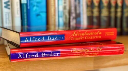 Two red Alfred Bader books, Adventures of a Chemist Collector and Chemistry & Art, lying on a shelf.
