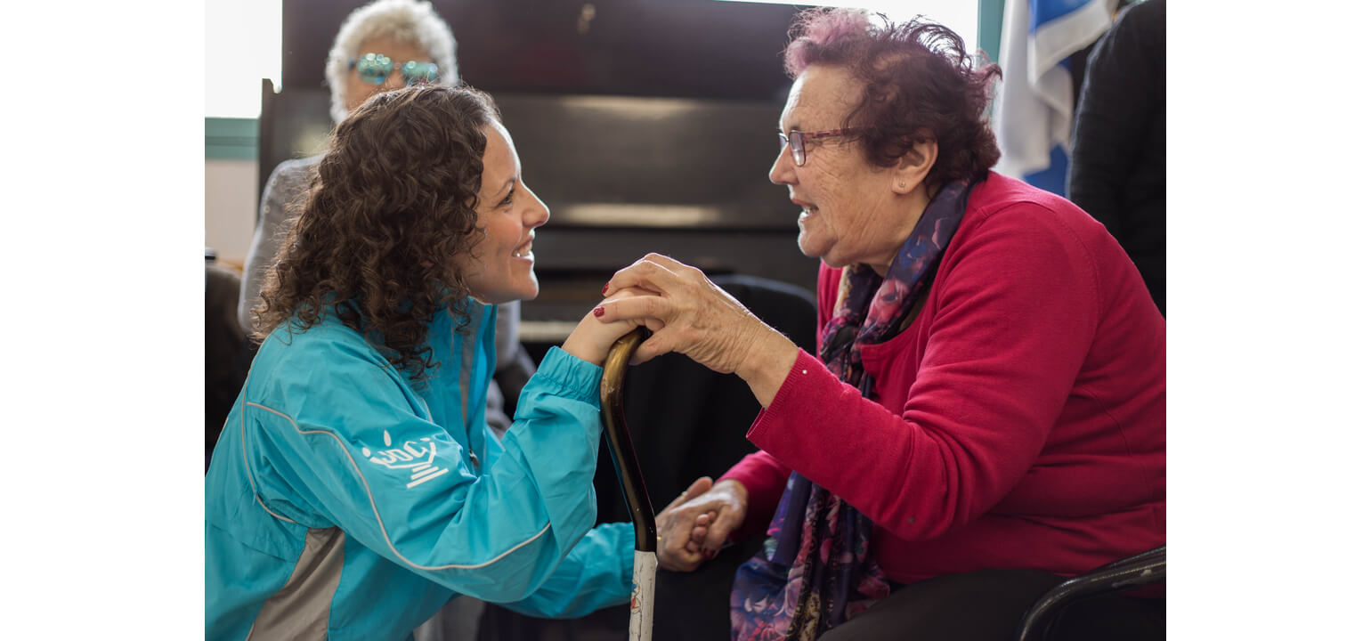 Smiling young woman in blue jacket gently holds the hand of an elderly woman with a cane, showing warm support.