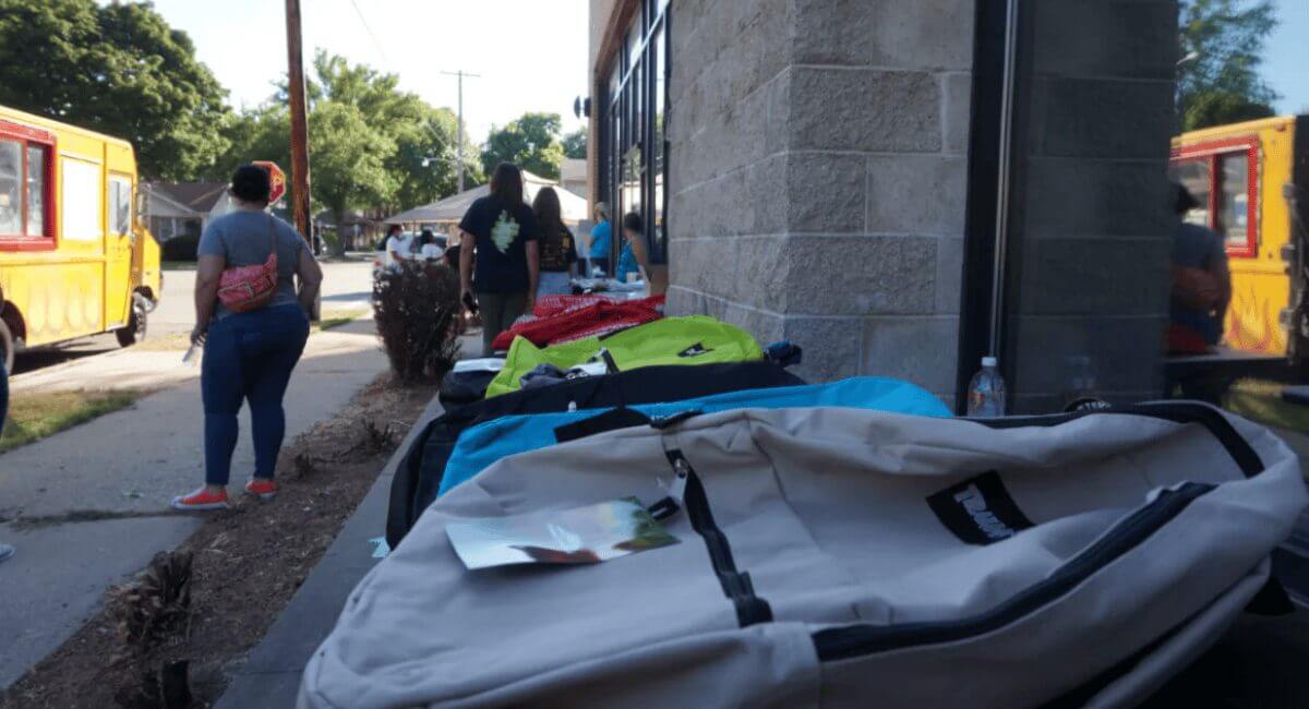Guests browse colorful backpacks at Chat with Bader March 2025 outdoor event. Yellow food trucks nearby.