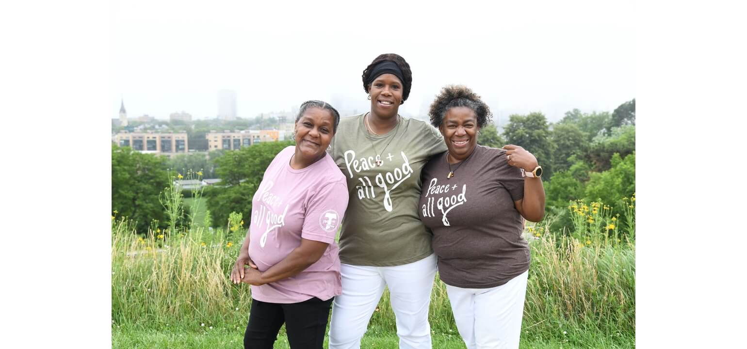 Smiling Black women from Clare community in 'Peace + all good' shirts, standing in a grassy field with a city backdrop.