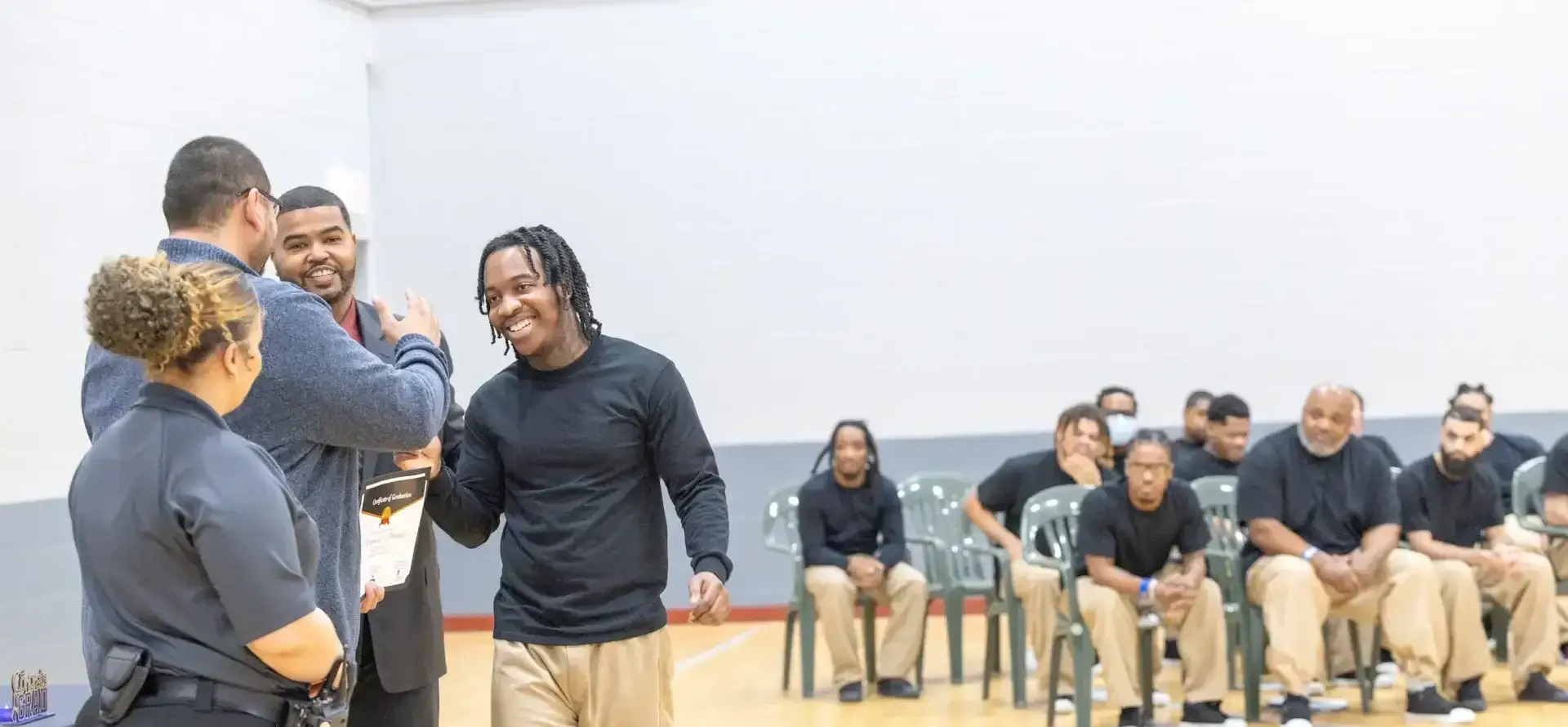 Smiling young man receives graduation certificate from mentor at CommunityMatters program. Diverse men watch.