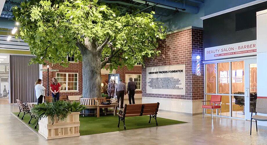 Large indoor park-like community space with an artificial tree, benches, a Green Bay Packers Foundation wall, and a barber shop.