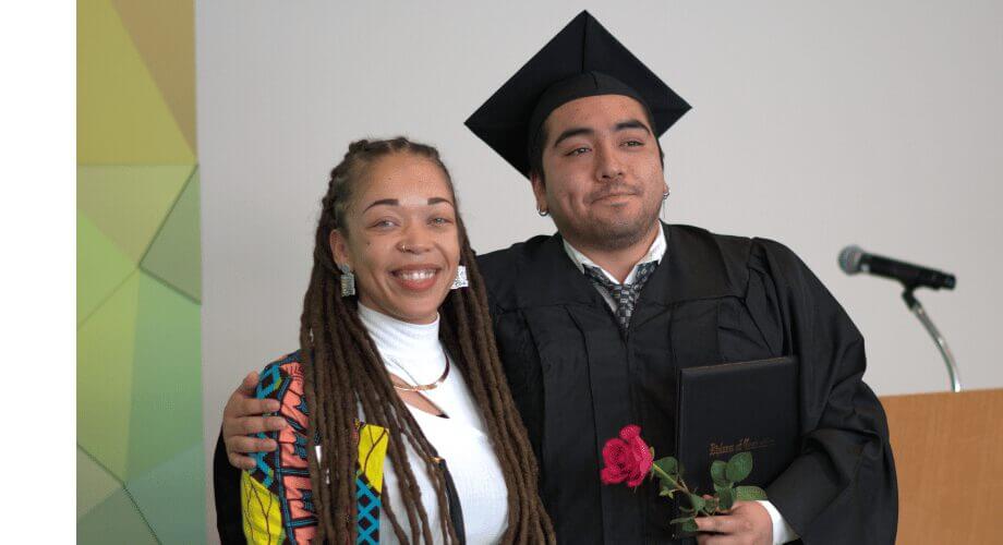 Proud GED graduate in cap and gown holding diploma and rose, celebrating with Joyce.