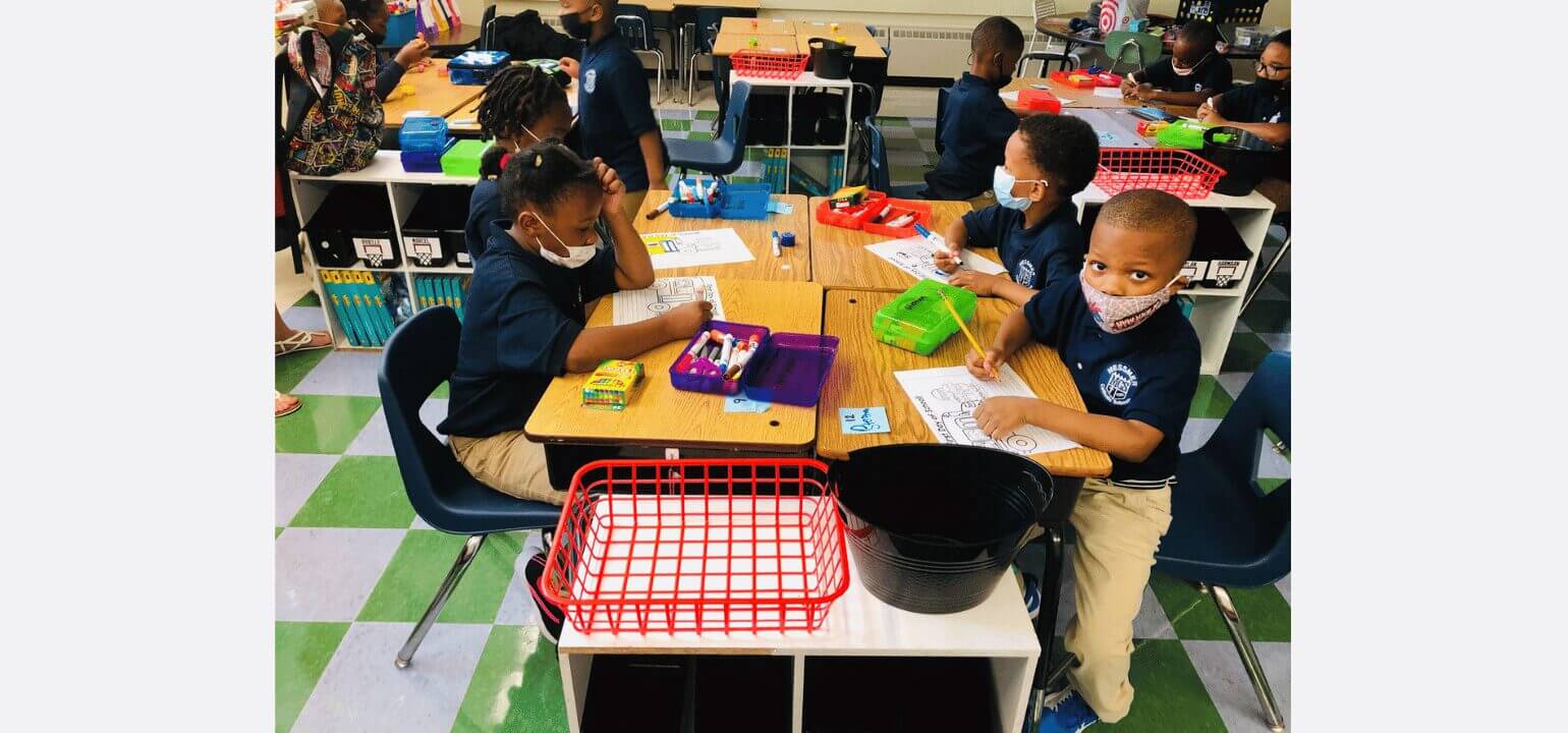 Young MSM students in uniform and masks sit at desks, engaged in art activities and coloring in a classroom.