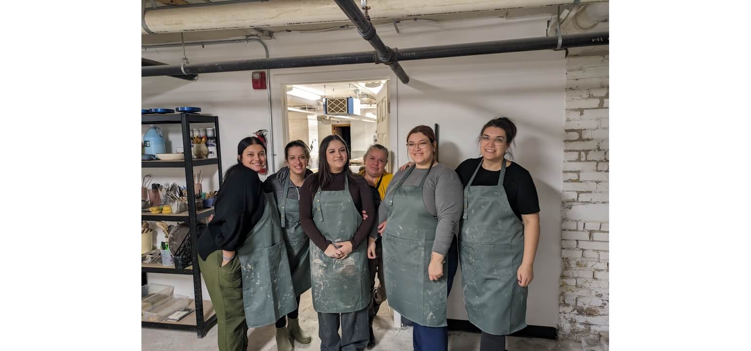 Six smiling women in clay-stained aprons pose in a pottery workshop, with art supplies on shelves nearby.