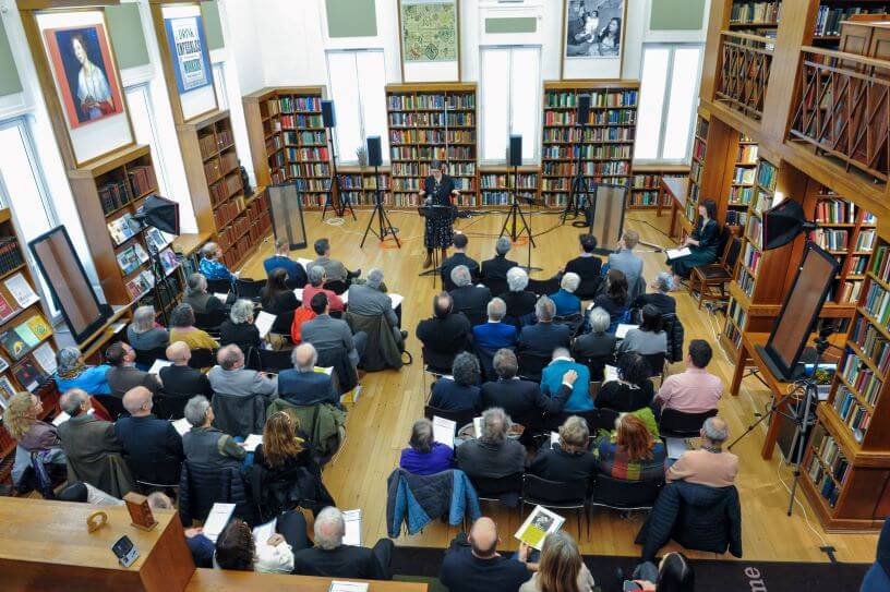Woman speaks to an audience at a podium during a library lecture, surrounded by packed bookshelves.
