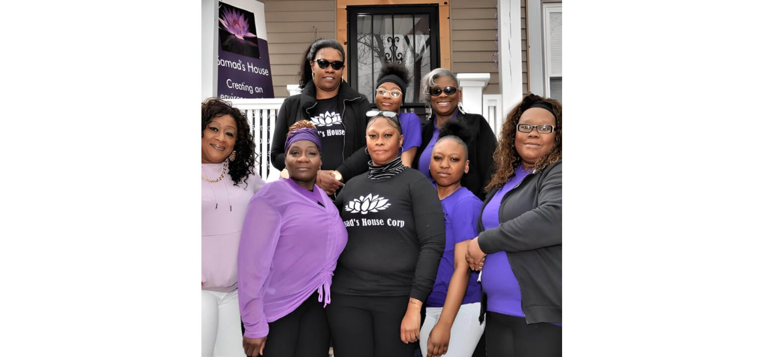 Samad’s House Corp Team outside their home Eight diverse women from Samad's House Corp in black and purple shirts smile outdoors in front of their building.
