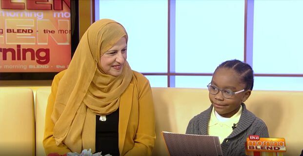 The Morning Blend: Woman in Hijab & Child Interview Woman in yellow hijab smiles at young child with glasses reading a book on The Morning Blend TV show set.