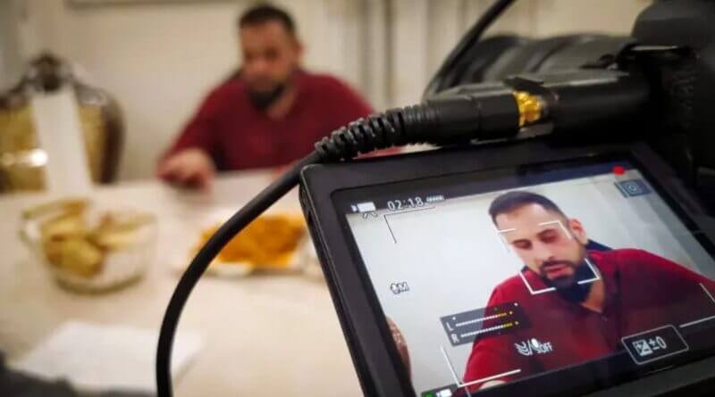 Man in red shirt recorded by professional video camera. Live feed on screen shows man's face with recording indicators, audio levels.