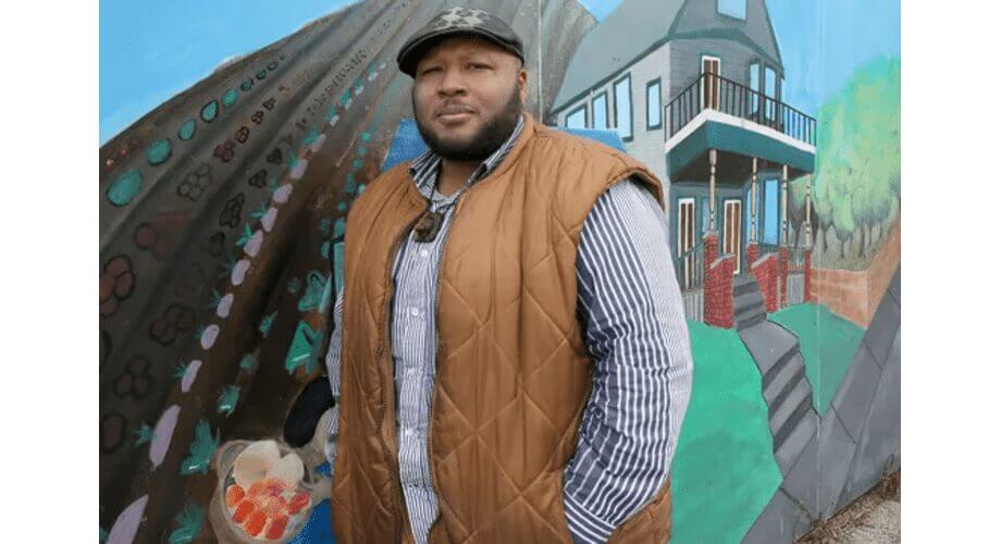 Black man with beard, wearing a brown quilted vest and flat cap, standing confidently in front of a vibrant street art mural.