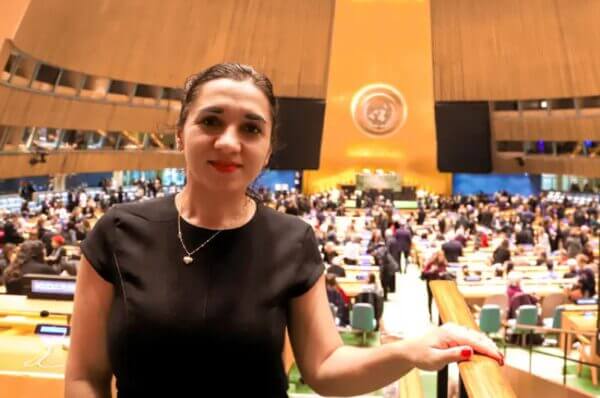 Professional woman in black top smiles, attending a UN General Assembly session. Delegates and emblem visible in the grand hall.