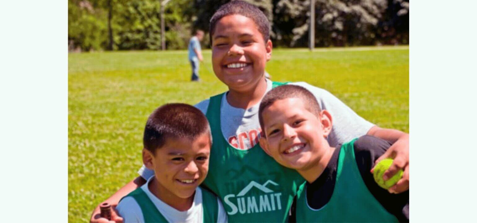 Summit Educational Program – Happy Kids Outdoor Activity Three smiling boys in green Summit jerseys on a sunny field, enjoying an educational outdoor activity or summer camp.