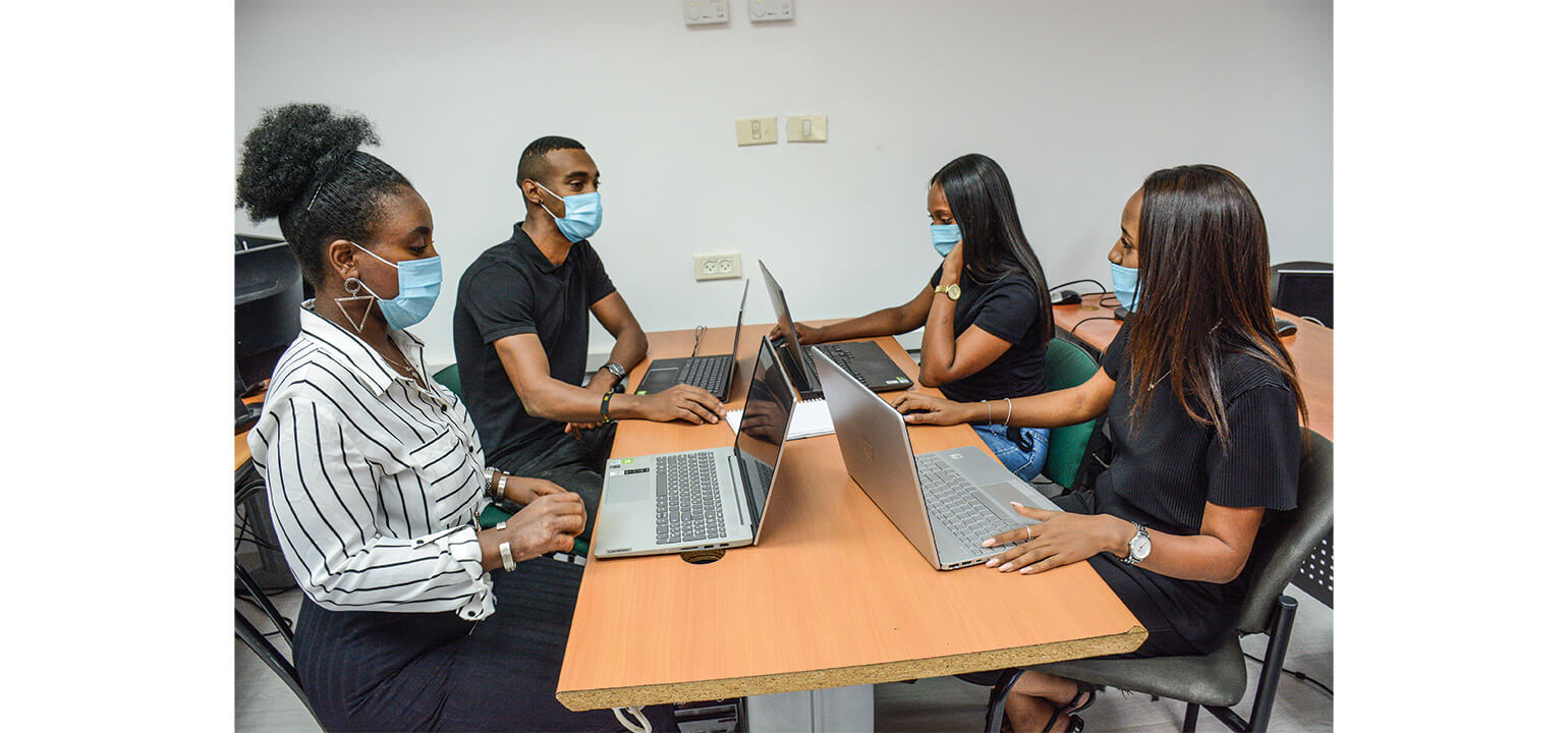 Diverse young adults in masks collaborating on laptops during a tech career training session.