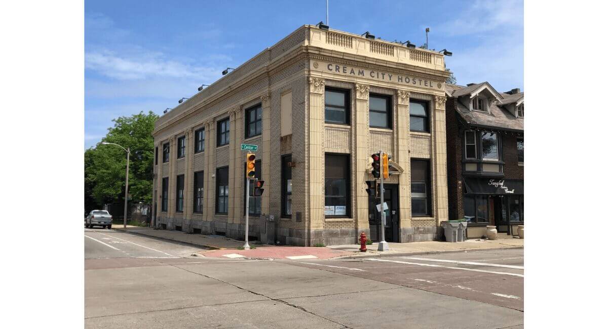 Cream City Hostel, a historic light brick building, at a sunny urban street corner with traffic lights and street signs.