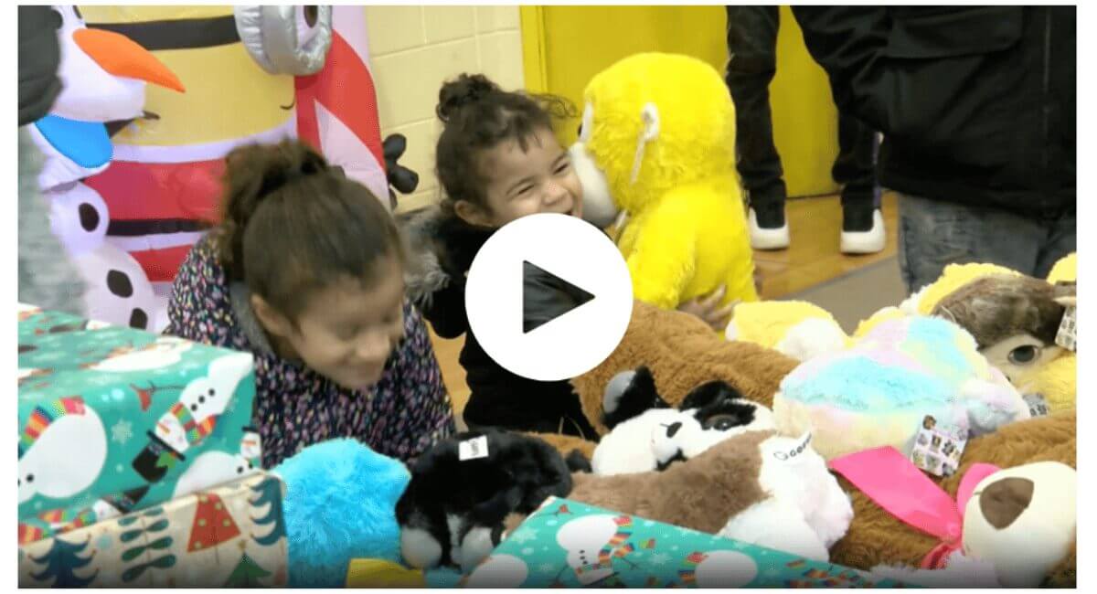 Kids Joyfully Receiving Holiday Gifts and Stuffed Animals Smiling child hugs a yellow plush toy, surrounded by wrapped holiday gifts and various stuffed animals at a festive event.
