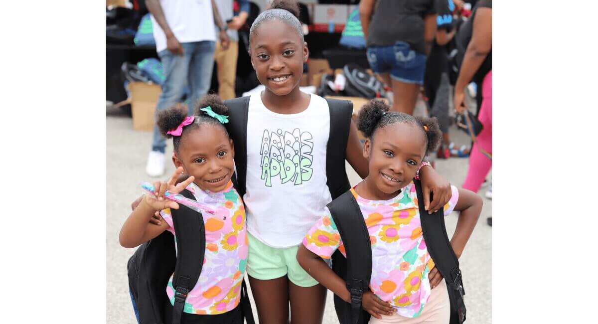 Three smiling Black girls with backpacks, excited for back to school or a community youth event.