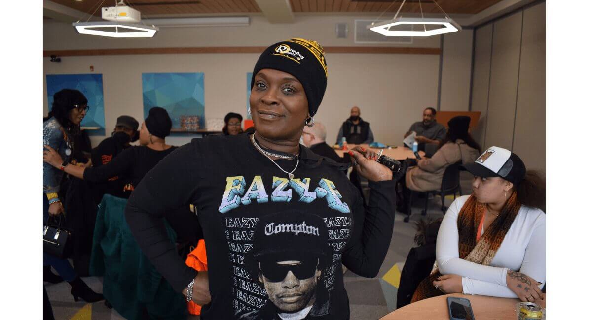 Confident woman in Eazy-E Compton t-shirt and beanie smiles, surrounded by people at a lively community gathering.