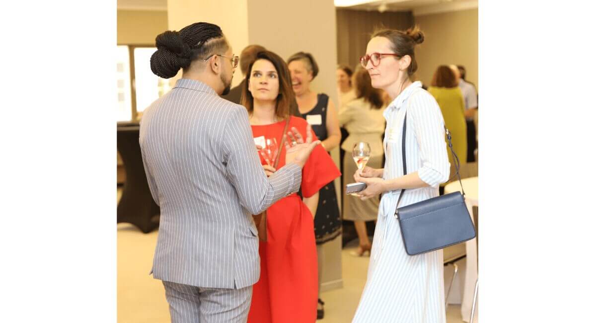 Man in striped suit speaks to two women at a professional networking event. Attendees mingle, one woman holds a wine glass.