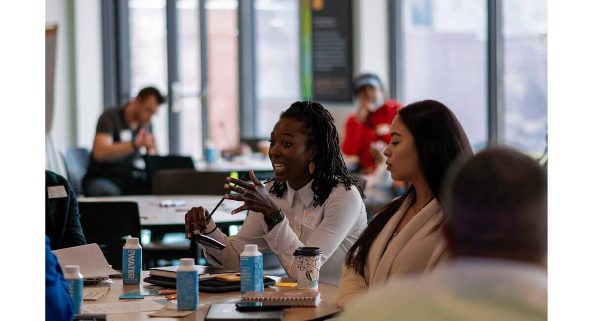 Professional Woman Speaking at Business Workshop Black woman with dreadlocks speaking animatedly at a business workshop table, gesturing with hands. Diverse colleagues listen.