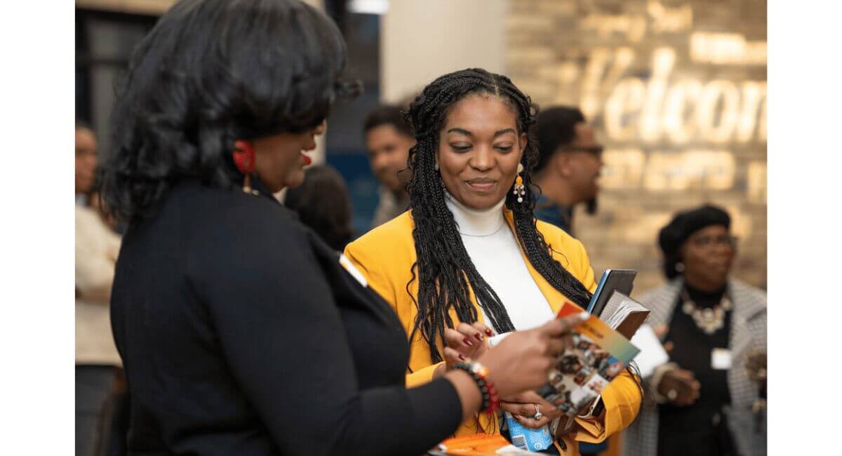 Two Black women networking at a professional event, one reviewing brochures. 'Welcome' sign visible in background.