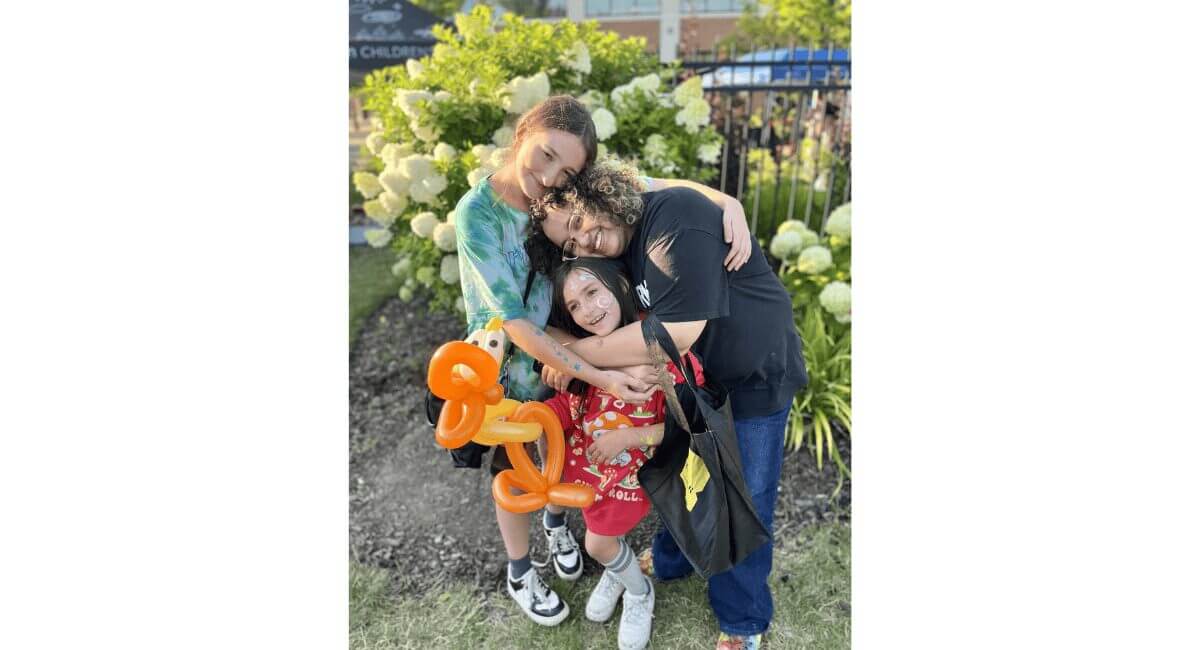 Two adults hugging a smiling child with face paint & a balloon animal at a vibrant outdoor children's event.