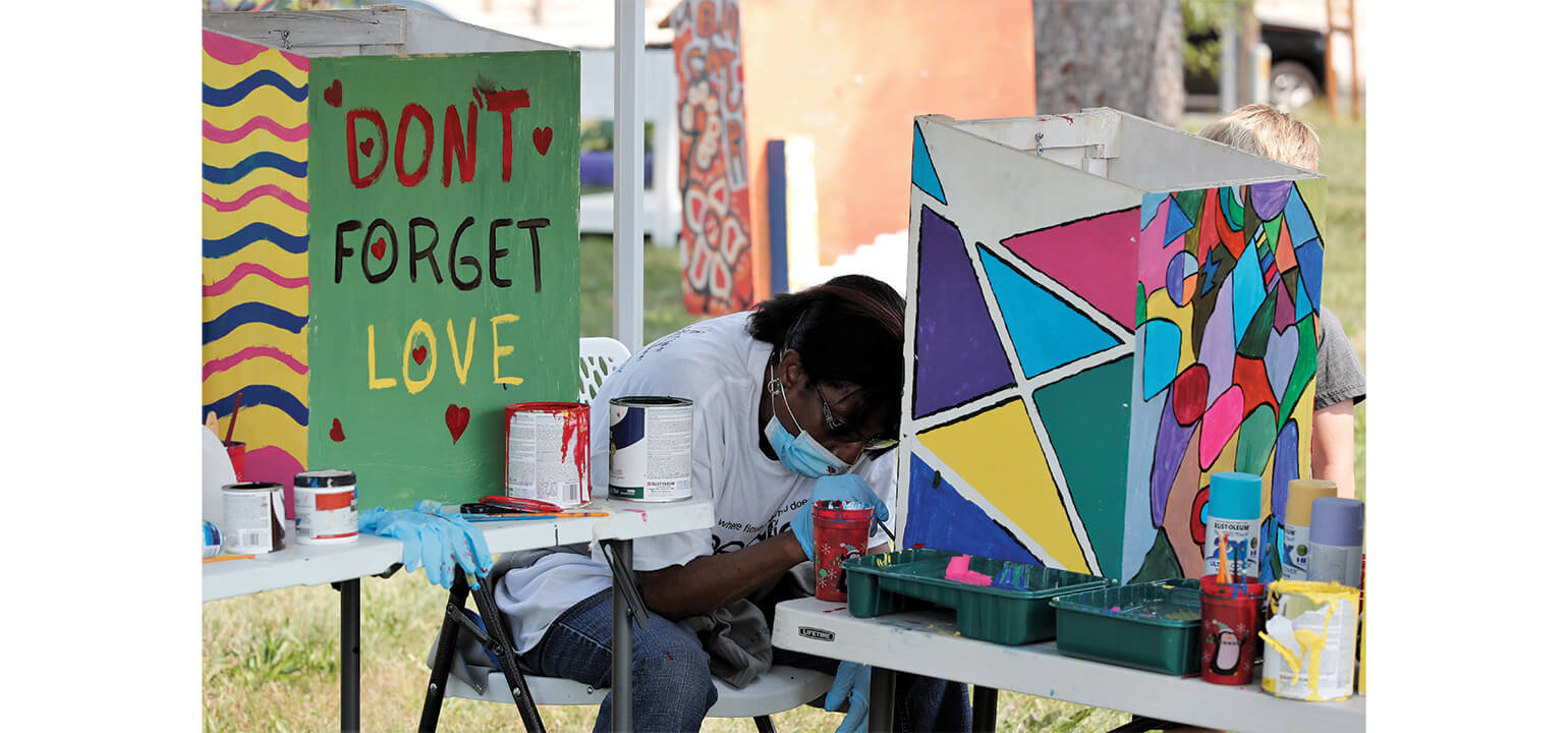Masked artist painting vibrant geometric designs on an art piece, next to a panel reading "Don't Forget Love".