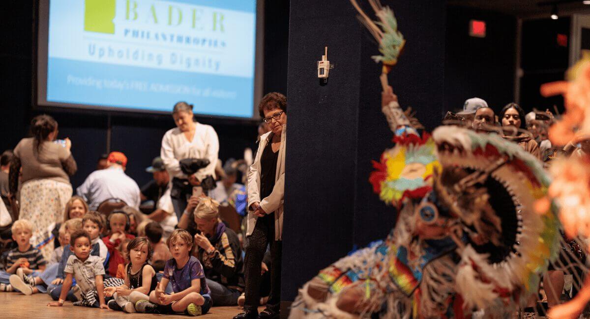 Bader Philanthropies banner behind children watching a vibrant Native American cultural dance performance.