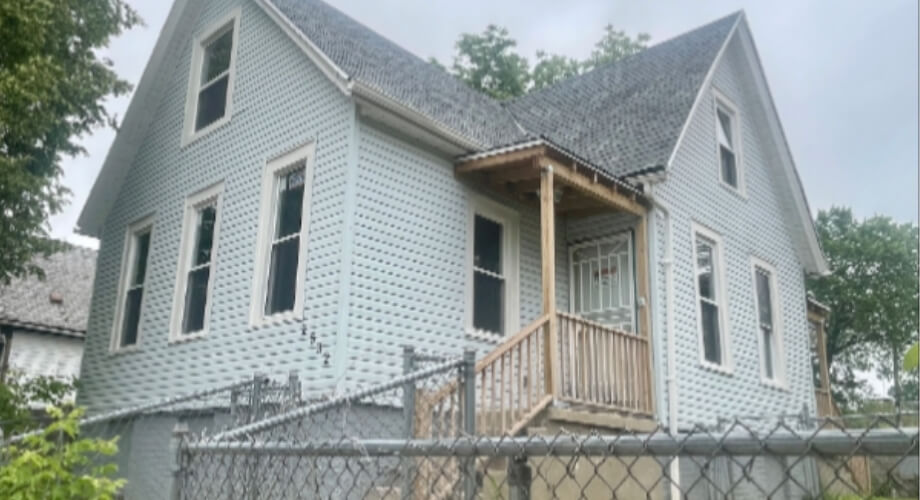 Old Residential House with Porch & Chain Link Fence Distressed two-story house with light blue siding, wooden porch, and gabled roof behind a chain-link fence.