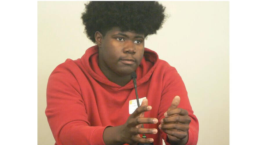 Young Black male in red hoodie speaks into a microphone, gesturing with hands during a dynamic presentation or panel discussion.