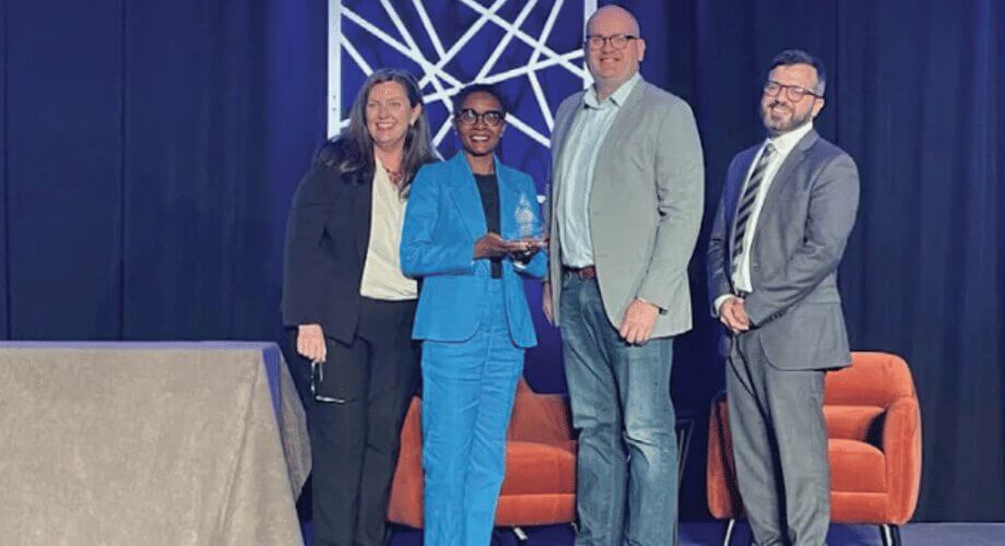 Diverse team of business leaders on stage, woman in blue suit proudly holds an award for achievement at an event.