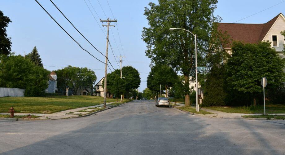 Quiet Residential Street View with Houses and Trees Quiet residential street lined with houses, mature trees, utility poles, and a parked car. Sunny neighborhood view.