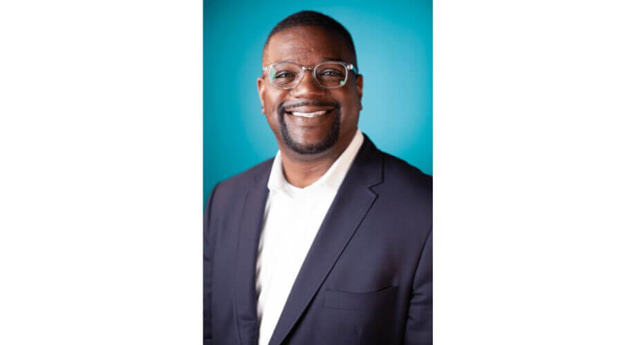 Professional Headshot: Smiling Man in Glasses and Suit Smiling Black man in clear glasses, dark suit, white shirt, and beard against a vibrant blue background.