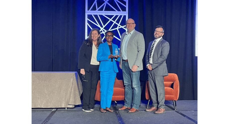 Four professionals on stage; woman in blue suit accepts glass award, flanked by colleagues at a corporate event.