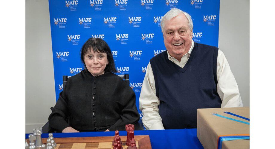 MATC Chess Event: Smiling Guests & Gift Smiling man and woman at MATC event, chessboard & gift. Background shows MATC logo & 'Transforming Lives'.