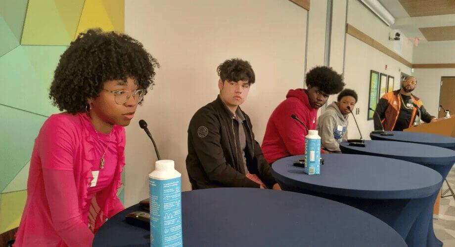 Diverse young professionals engaging in a panel discussion, seated with microphones. A speaker stands at a podium.